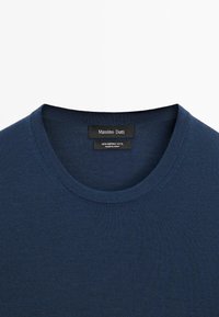 Maglione in lana merino blu navy con texture a coste e scollo rotondo. L'etichetta mostra il nome del marchio e i dettagli del tessuto sulla parte interna del collo.