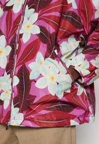 Floral jacket with a shiny, lightweight fabric. Features large white flowers and red leaves on a pink background. Includes pockets and elastic cuffs.