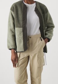 Veste en polaire verte avec des accents marron, fermeture éclair et manches longues, associée à un pantalon cargo beige comportant des poches latérales et un détail de logo.
