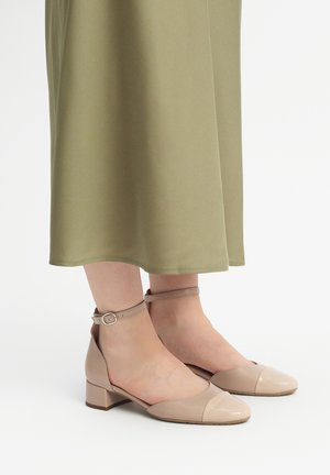 Beige lakatut nahkasaappaat nilkkaremmillä, joissa on neliömäinen kärki ja tukevat korot, yhdistettyinä oliivinvihreään mekkoon, joka ulottuu polven alapuolelle.