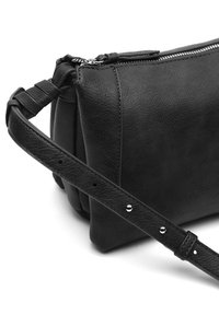 Crna kožna torbica preko tijela s teksturiranim završetkom, zatvaranjem na zip i podesivim remenom s metalnim zakovicama. Pravokutnog oblika s zaobljenim rubovima.