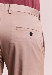 Pantaloni beige con una superficie liscia, dotati di una tasca posteriore con un bottone nero e un design a taglio dritto.