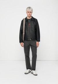 Black leather jacket with zip closure, gray cargo pants, and white sneakers with black stripes. Gray shoulder bag rests on one shoulder.