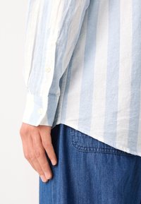 Chemise en lin rayée bleue et blanche avec des poignets à boutons ; ourlet arrière plus long ; associée à un tissu en denim, montrant une vue latérale de la chemise.
