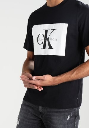 Mann trägt ein schwarzes Calvin Klein Jeans T-Shirt mit weißem Logorechteck, die Hände verschränkt, kombiniert mit abgenutzter dunkelgrauer Jeans.