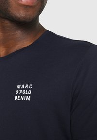 Navy sininen lyhythihainen t-paita, joka on valmistettu sileästä puuvillasta ja jossa on valkoinen painettu logo "MARC O'POLO DENIM" vasemmassa rintataskussa.