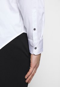 Vit skjorta med lång ärm, med svarta knappar och skräddarsydd passform. Nederkanten är något asymmetrisk, med en slät tygstruktur.