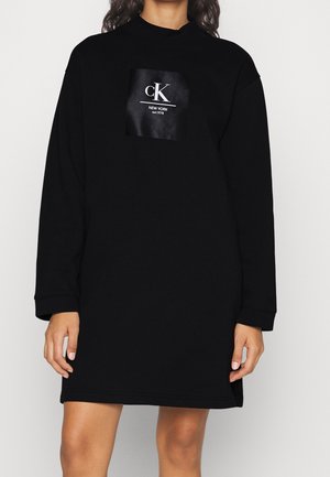 Kobieta w długiej czarnej sukience dresowej z białym logo „cK New York est. 1978” na piersi, stojąca na jednolitym białym tle.