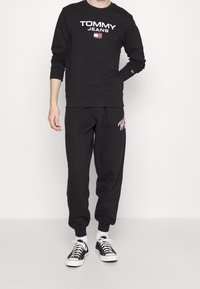 Sudadera negra con el logo blanco de "TOMMY JEANS", cuello redondo y mangas largas. Pantalones de chándal negros con detalle de logo rosa, ajustados en los tobillos.