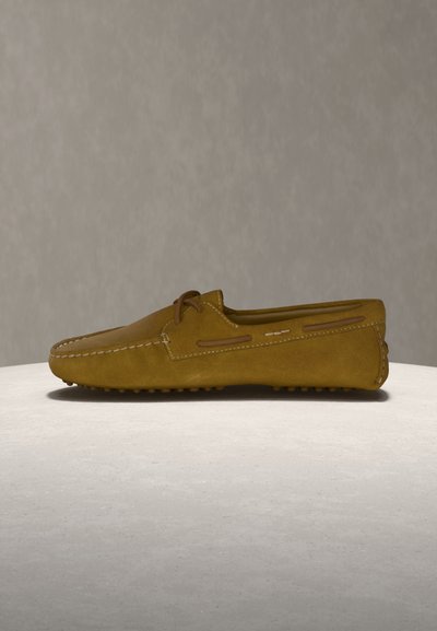 Καφέ μοκασίνι σουέντ με εμφανείς ραφές, λεπτομέρεια κορδονιών και υφή στη σόλα, τοποθετημένο σε ανοιχτό γκρι επιφάνεια με απλό φόντο.