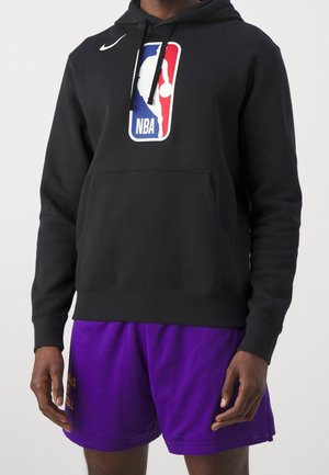 Mann trägt schwarzen NBA-Hoodie mit Fronttasche und lila Basketballshorts vor schlichter Hintergrund.