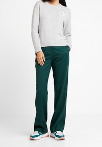 Grauer Strickpullover mit einem CK-Logo, kombiniert mit locker sitzenden dunkelgrünen Hosen und mehrfarbigen Laufschuhen.