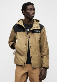 Το μπουφάν σε χακί και μαύρο διαθέτει αδιάβροχο υλικό, κουκούλα και κούμπωμα με τρουκ. Φοριέται πάνω από μαύρη μπλούζα και μπεζ παντελόνι.