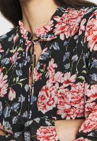 Blouse noire à fleurs avec de grandes fleurs roses et bleues, un col à volants, une fermeture à nouer et des manches longues plissées. Texture de tissu doux.