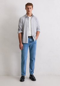 Szara i biała pasiasta koszula na guziki na białej koszulce, dobrana do niebieskich dżinsów i czarnych butów. Model stoi swobodnie, ręce w kieszeniach.