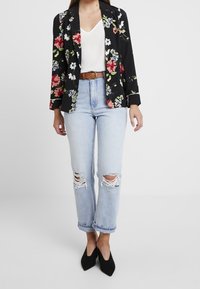 Blazer med blommönster i svart, långärmad, kombinerad med en vit topp, ljusblå rivna jeans och svarta stilettklackar.