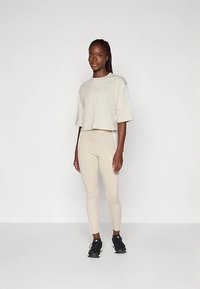 Γυναίκα στέκεται μπροστά σε λευκό φόντο φορώντας μπεζ χαλαρή μπλούζα και αντίστοιχες κολάν με μαύρα sneakers, κοιτάζοντας μπροστά με μια ελαφριά χαμόγελο.