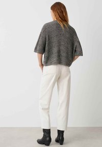 Fülliger Kurzarmpullover aus grauem Strickstoff, kombiniert mit weißen Hosen und schwarzen Stiefeletten, von hinten betrachtet.