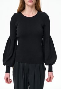 Kobieta w czarnym prążkowanym swetrze z bufkami na rękawach i czarnych plisowanych spodniach, stojąca na białym tle.