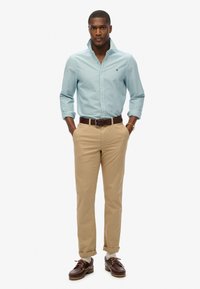 Camisa de botones a rayas azul claro, pantalones chinos beige, zapatos de barco de cuero marrón. Estilo casual con mangas dobladas y cinturón marrón oscuro.
