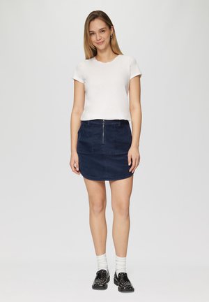 Witte T-shirt met ronde hals, gecombineerd met een marineblauwe corduroy minirok met een rits aan de voorkant en zakken. Zwarte gespte schoenen en witte sokken.