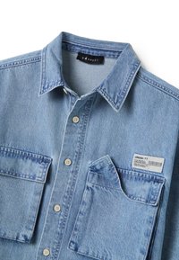 Camisa de denim azul claro con cuello, dos bolsillos frontales, cierre de botones y una etiqueta en el bolsillo derecho que muestra información de estilo y tamaño.