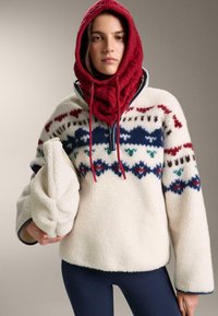 Fleecetröja i krämfärg med blå, röda och gröna mönstrade detaljer. Bärs med en röd stickad huva. Håller en krämfärgad påse. Tajta byxor syns.