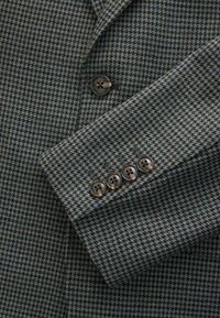 Textured blue and brown houndstooth patterned fabric of a jacket sleeve, featuring five dark button accents on the cuff.