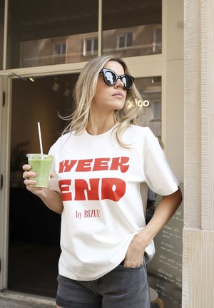 Witte t-shirt met gedurfde rode tekst "WEEK END by BIZUU," gecombineerd met donkere shorts. Houdt een groene drank vast in een doorzichtige beker. Zwarte zonnebril op.