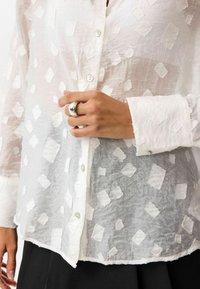 Transparante witte blouse met knopen, voorzien van textuur met diamantvormige lappen, geknoopte mouwen en een hand met een grote zilveren ring die nabij de knopen rust.