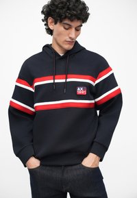 Sudadera color azul marino con rayas horizontales rojas y blancas, con un pequeño parche de logo. Tejido suave, capucha con cordón y corte relajado.