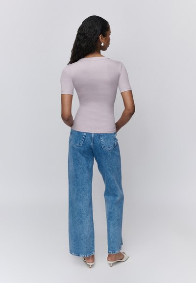 Femme aux longs cheveux bouclés, portant un top moulant violet clair à manches courtes, un jean bleu à jambes larges, des sandales blanches à talons, debout de dos.