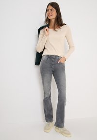 Haut beige à manches longues associé à un jean gris délavé. Le modèle se tient avec confiance, une veste verte drapée sur une épaule, portant des baskets claires.