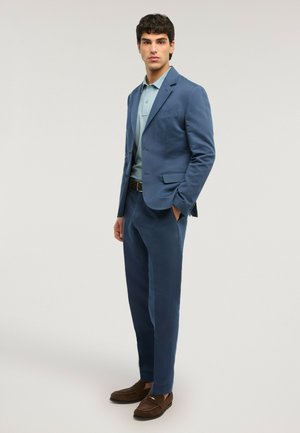 Jeune homme debout, les mains dans les poches, portant un costume bleu, une chemise bleu clair, une ceinture marron et des mocassins marron sur un fond uni.