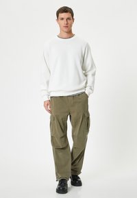 Witte sweatshirt met een ronde hals over een zwart-wit gestreept shirt, gecombineerd met olijfgroene cargobroek met zijzakken en zwarte schoenen.