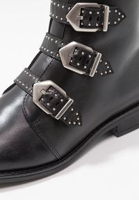 Černé kožené kotníkové boty s třemi kovovými přezkami a hroty. Hladký povrch s nízkým blokovým podpatkem.