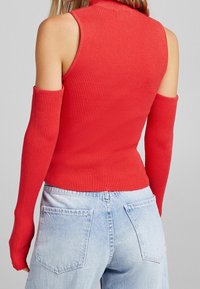 Röd ribbad ärmlös topp med hög krage och förlängda ärmar, matchad med ljusblå denimjeans med bakfickor.