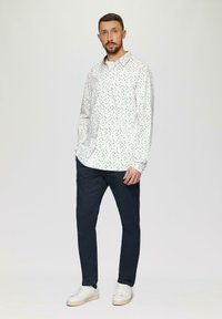 Witte shirt met gekleurde stippen, lange mouwen en een button-down kraag, gecombineerd met donkerblauwe broek en witte sneakers.