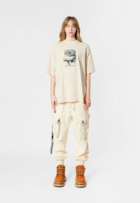 Camiseta oversize beige con estampado gráfico en el centro, combinada con pantalones cargo crema con bolsillos con cremallera y botas marrones con cordones.