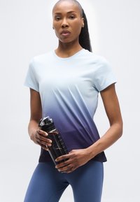 Γυναίκα με μπλουζάκι γυμναστικής σε βαθμίδες μπλε χρώματος και μπλε κολάν κρατώντας ένα μαύρο παγούρι νερού σε απλό φωτεινό φόντο.