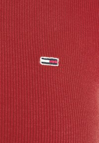Roter gerippter Stoff mit einem kleinen rechteckigen Logopatch, der horizontal gestreift in Weiß, Marineblau und Rot in der Mitte des Textils angebracht ist.