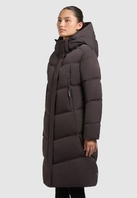 Abrigo largo acolchado en marrón oscuro con un cuello alto y capucha. Presenta costuras verticales y un bolsillo con cremallera en el lado izquierdo.