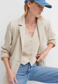 Blazer di lino beige con maniche arrotolate, indossato sopra un gilet abbinato con tre bottoni, abbinato a jeans di denim blu.