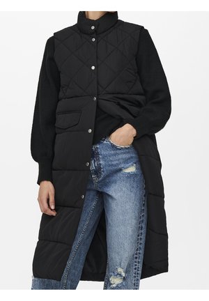 Long gilet matelassé noir avec boutons-pression, porté sur un pull noir et un jean bleu déchiré, avec une main dans la poche.