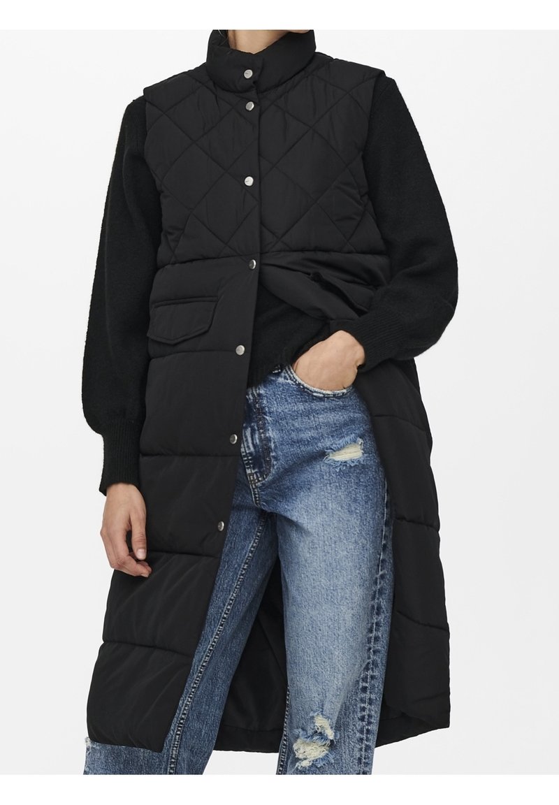 Long gilet matelassé noir avec boutons-pression, porté sur un pull noir et un jean bleu déchiré, avec une main dans la poche.