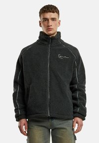 Tumehall fleece-saal koos kõrge kraega, ees lukuga ja valgete õmblusaktsentidega. Omab pehmet, tekstuurset välimust.