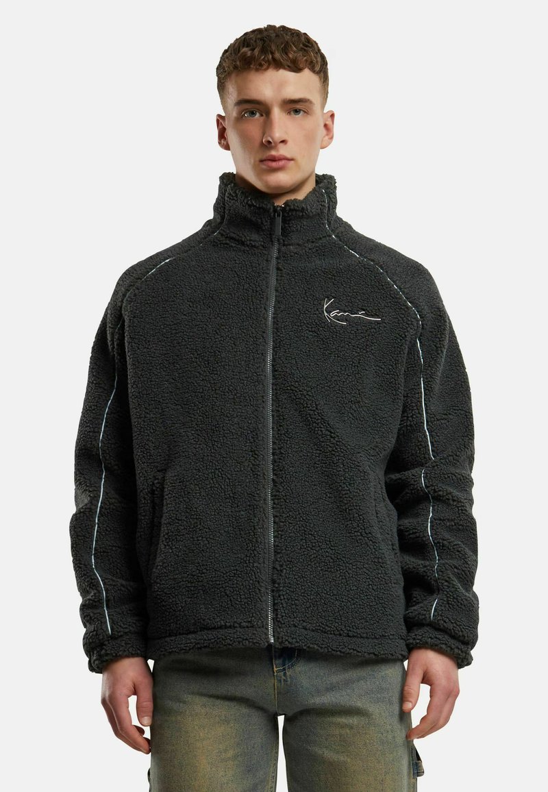 Tumehall fleece-saal koos kõrge kraega, ees lukuga ja valgete õmblusaktsentidega. Omab pehmet, tekstuurset välimust.