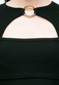 Top nero a coste con un accento di hardware dorato rotondo sul collo, dotato di un design con taglio e tessuto strutturato.