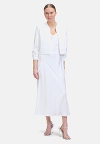 Robe maxi blanche sans manches avec un décolleté en V, associée à une veste blanche courte et des sandales à talons hauts argentées. Tissu lisse et fluide.