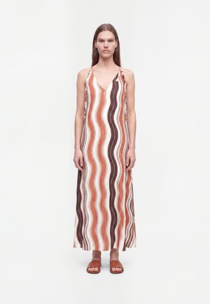 Femme debout portant une robe maxi sans manches à rayures ondulées verticales en marron, rouille et beige, assortie à des sandales plates marron.